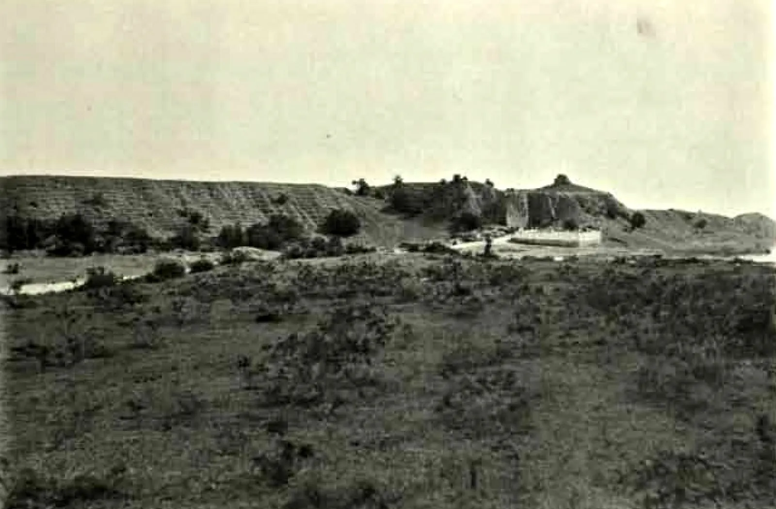 A long, low earthen rampart covered with scrub, seen across an open grassy plain.