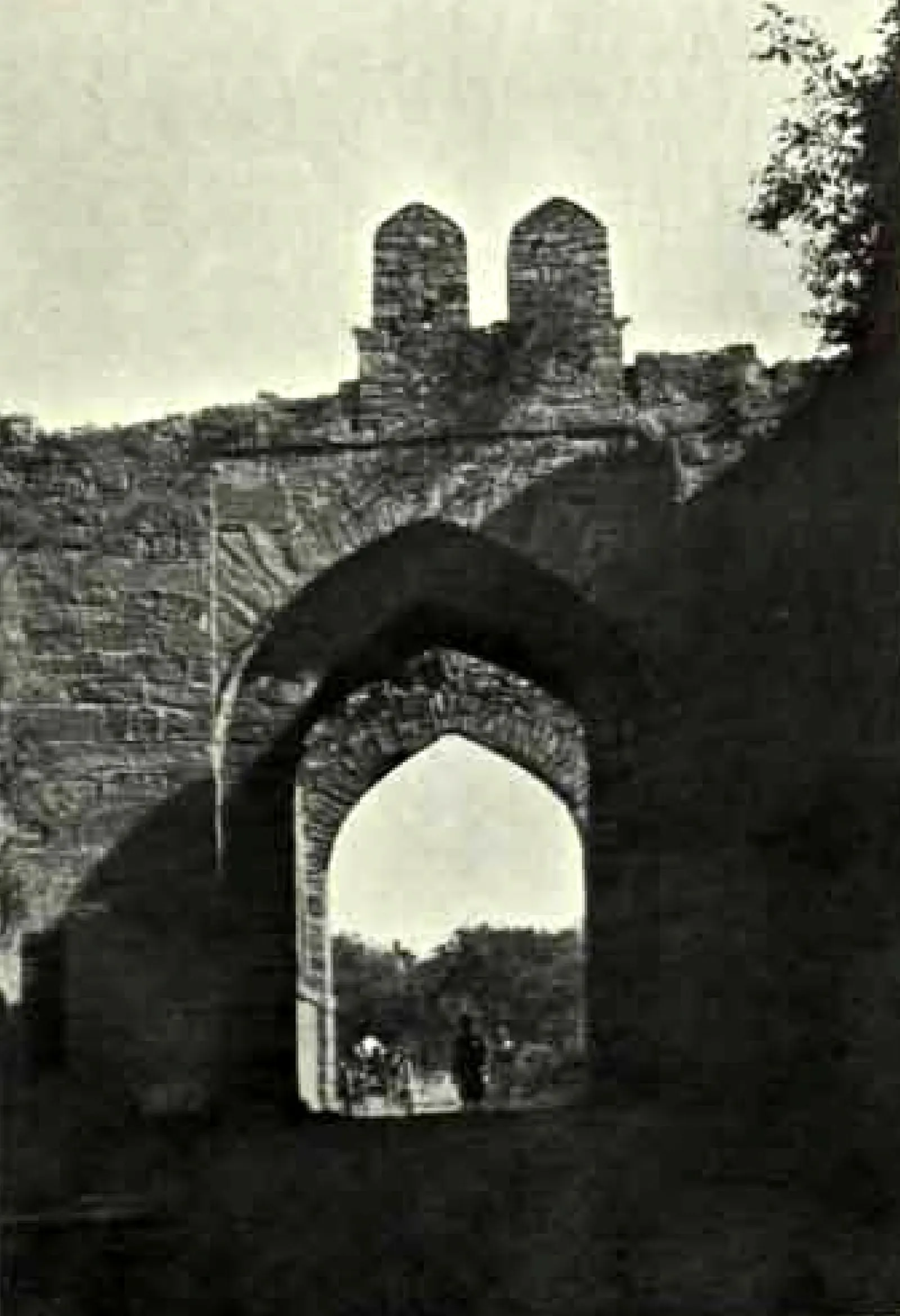 A pointed archway through a thick stone wall topped by two small turrets, with people in the bright opening.
