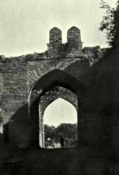 A pointed archway through a thick stone wall topped by two small turrets, with people in the bright opening.