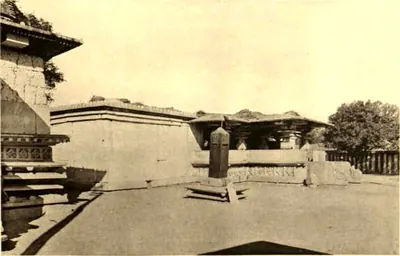 A paved courtyard with a small pillar on a base in front of a low flat-roofed temple, and the corner of another temple at the left.