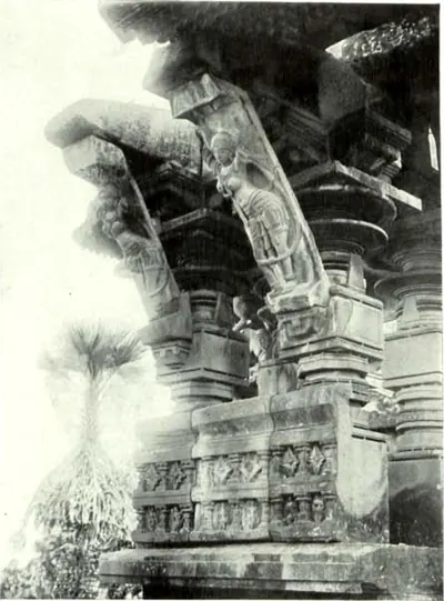Slanting stone brackets carved with standing female figures above pillar capitals, seen from below against the sky, with a palm beyond.