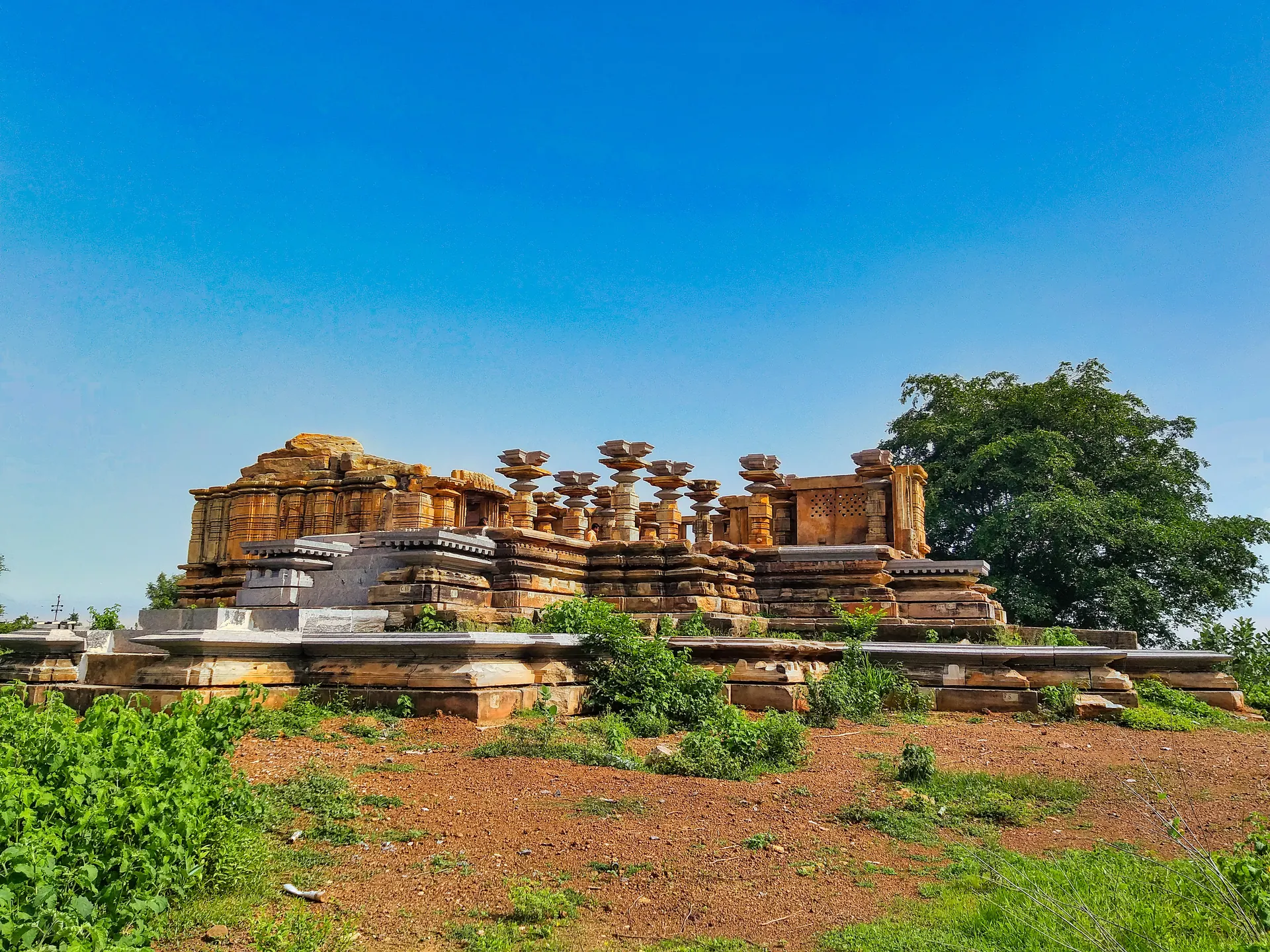 A stone temple on a raised platform in open red ground, its shrine walls partly rebuilt and a row of unroofed pillars in front, with a large tree at the right.