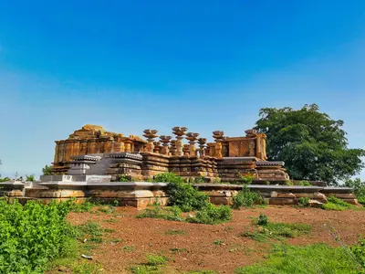 A stone temple on a raised platform in open red ground, its shrine walls partly rebuilt and a row of unroofed pillars in front, with a large tree at the right.