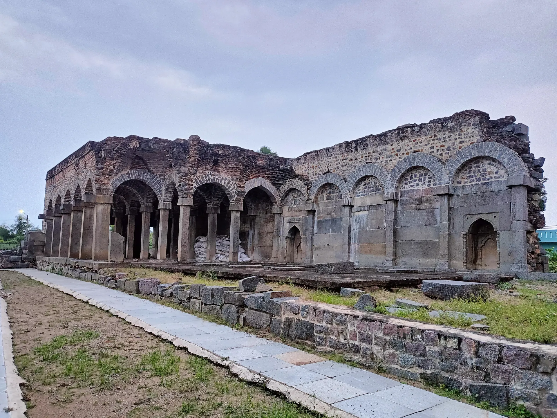 The partly ruined Konda Masjid: a long arcade of pointed arches on stone piers with collapsed upper walls, behind a paved path and a low rubble wall.