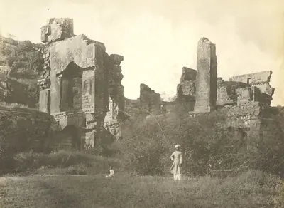 Roofless ruined walls and doorways of the upper storey of the palace, overgrown with plants, with a man standing in the foreground.
