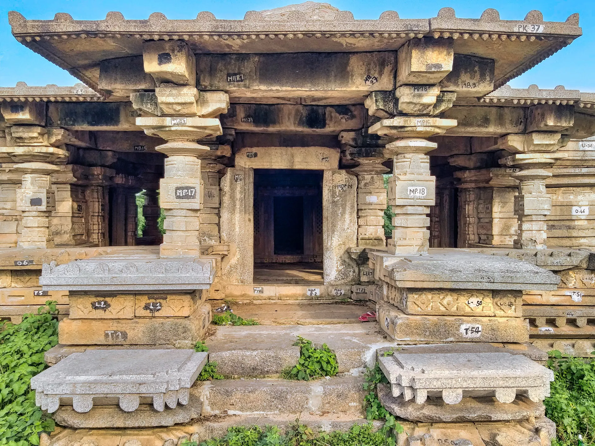 A pillared stone hall with a flat roof and wide eaves on a platform, steps leading to a central doorway, many stones painted with numbers.