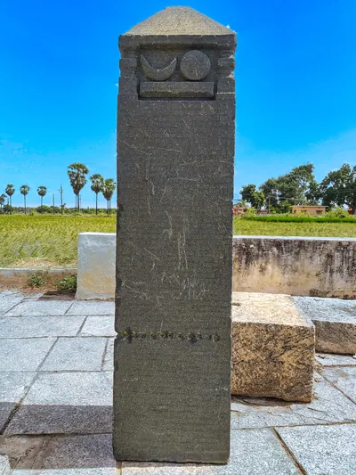 A square stone pillar inscribed on its faces, topped by a carved crescent moon and sun and a pyramidal cap, standing on a paved platform.