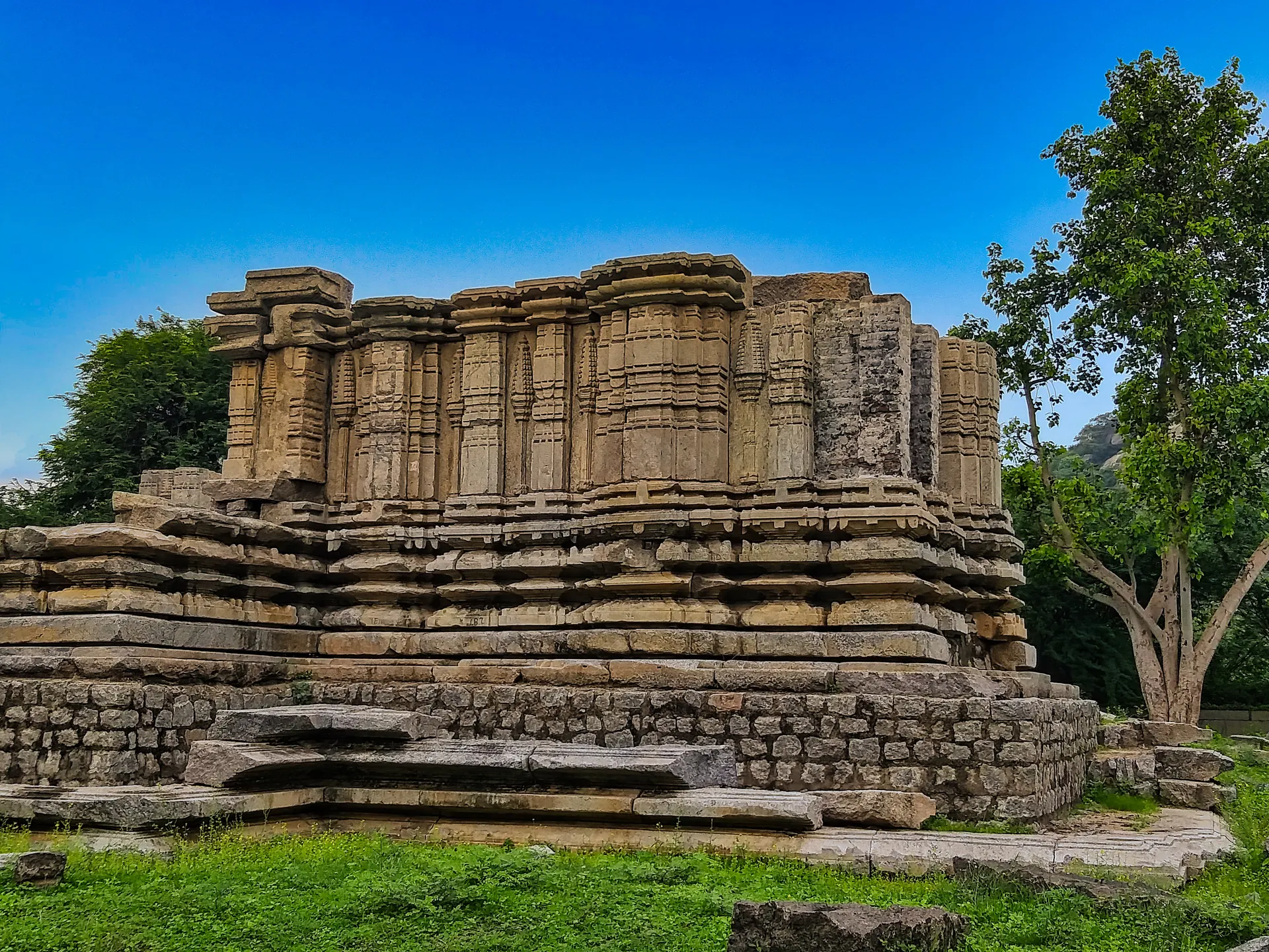 Ruined walls of a stone temple standing on a moulded base, the superstructure lost, beside a tree under a blue sky.