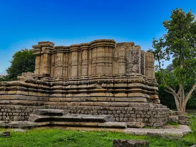 Ruined walls of a stone temple standing on a moulded base, the superstructure lost, beside a tree under a blue sky.