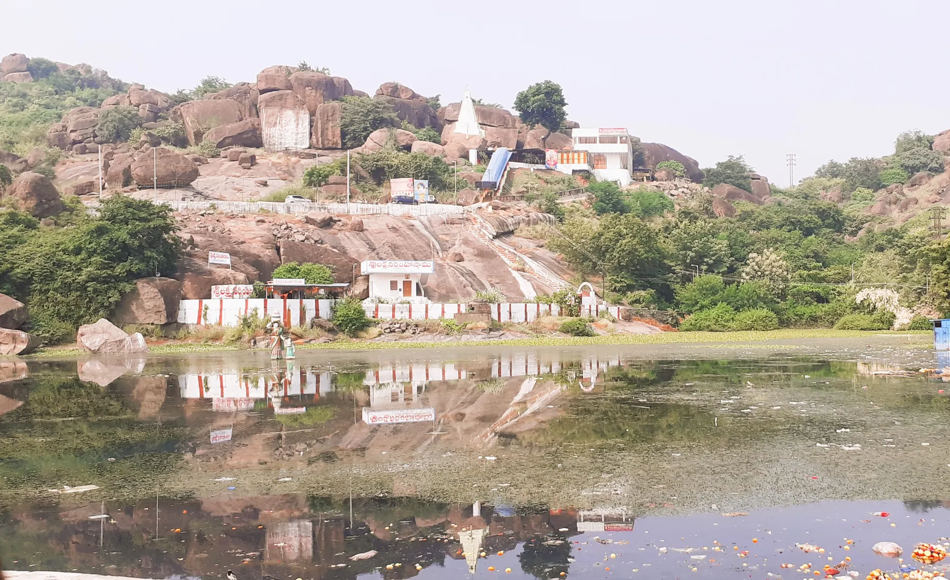 A rocky granite hill with a small white pyramidal temple tower near the top and white buildings on the slope, reflected in a pond in the foreground.