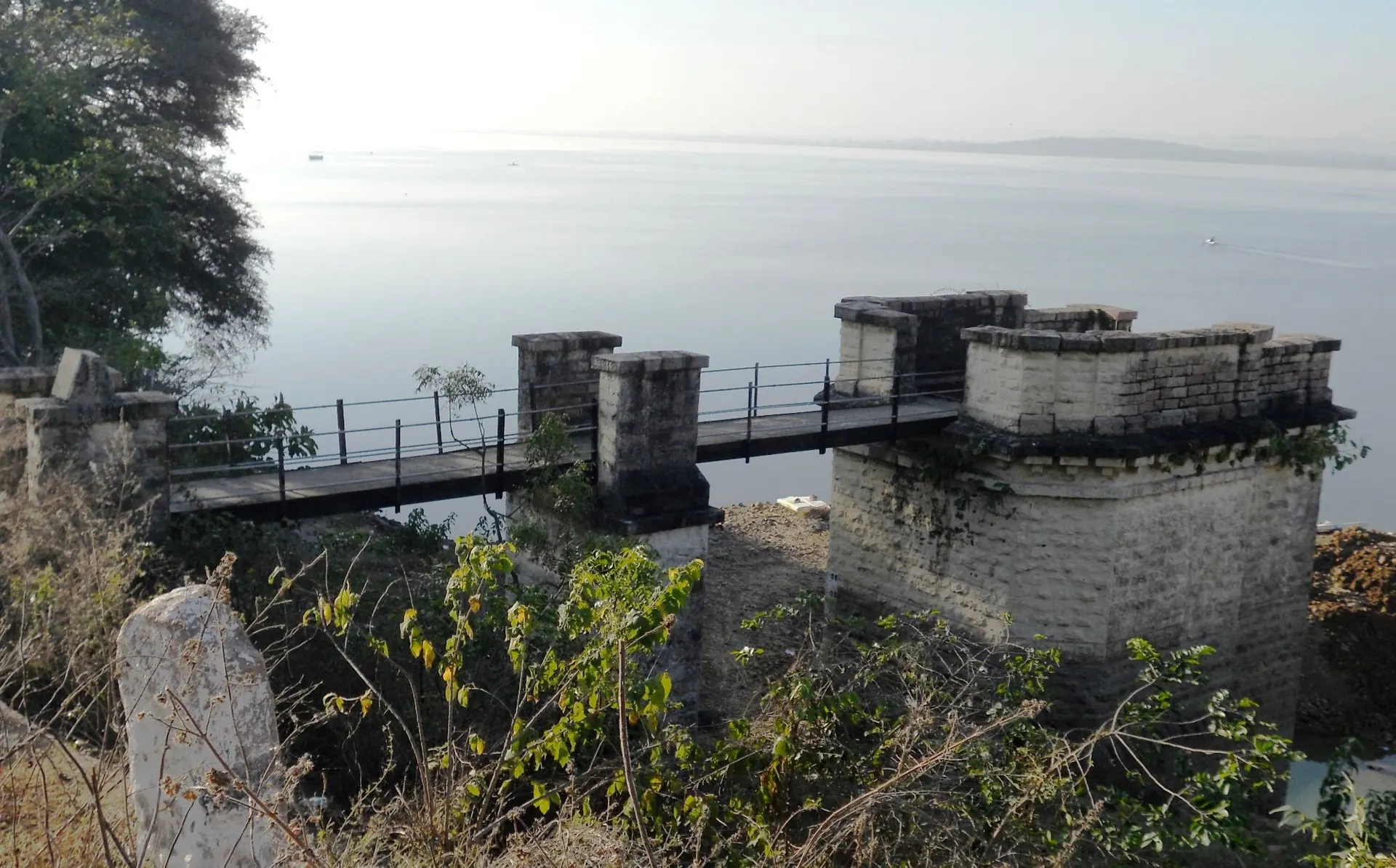 A square masonry tower with a notched parapet at the edge of the lake, linked to the bank by a narrow metal footbridge on masonry piers, with open water behind.