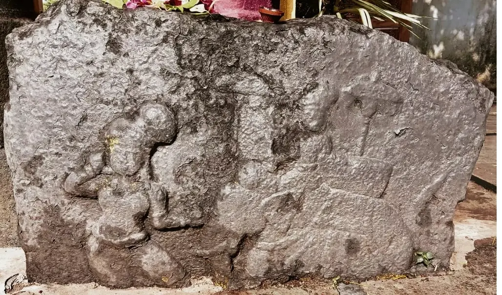 A weathered slab of dark stone with worn low-relief carvings, one apparently a kneeling person, with flowers lying on top.