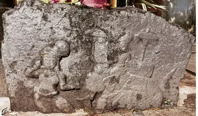 A weathered slab of dark stone with worn low-relief carvings, one apparently a kneeling person, with flowers lying on top.