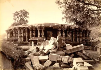 The pillared hall of the Thousand Pillar Temple behind a heap of fallen beams and pillar parts, a stone Nandi on the rubble and thatched huts at the left.