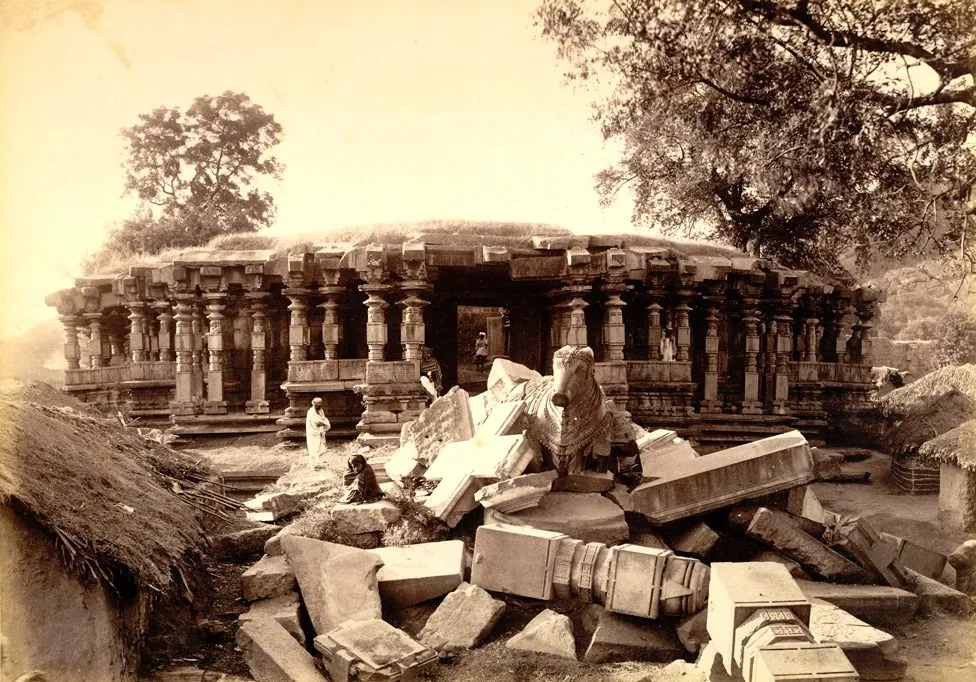The pillared hall of the Thousand Pillar Temple behind a heap of fallen beams and pillar parts, a stone Nandi on the rubble and thatched huts at the left.
