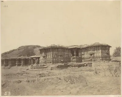 The square shrines of the Thousand Pillar Temple in front of a rocky hill, a long pillared hall extending to the left and mounds of earth and scrub in front.