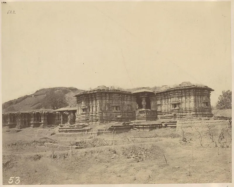 The square shrines of the Thousand Pillar Temple in front of a rocky hill, a long pillared hall extending to the left and mounds of earth and scrub in front.