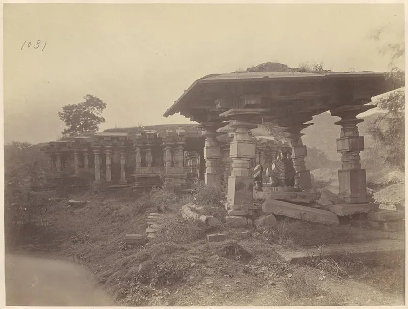 A stone pavilion with a heavy flat roof on carved pillars, standing on uneven ground with a long pillared building behind it at the left and loose stones in front.