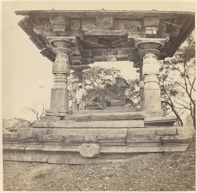 A carved stone bull under a flat stone roof held up by round carved pillars on a stepped platform, with a man standing beside the bull.