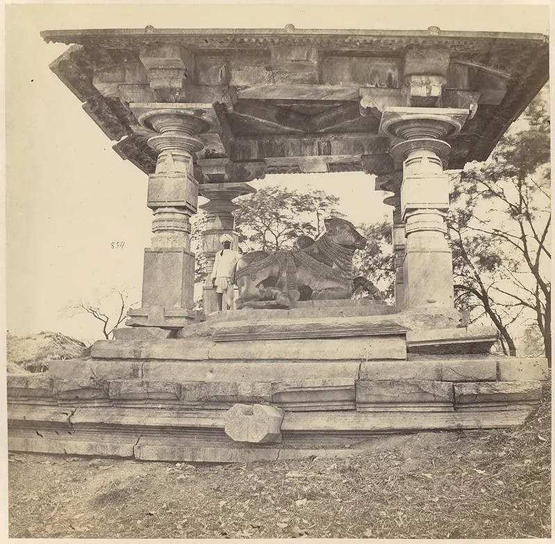 A carved stone bull under a flat stone roof held up by round carved pillars on a stepped platform, with a man standing beside the bull.