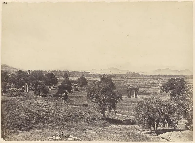 A wide view from a rocky hill over open fields and scattered trees, with stone gateways and pillars standing alone in the fields and low hills on the horizon.