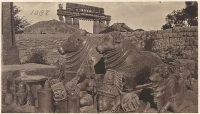 Two large carved stone bulls lying side by side among broken carved stones, a rough wall behind them and the top of a stone gateway rising above it.