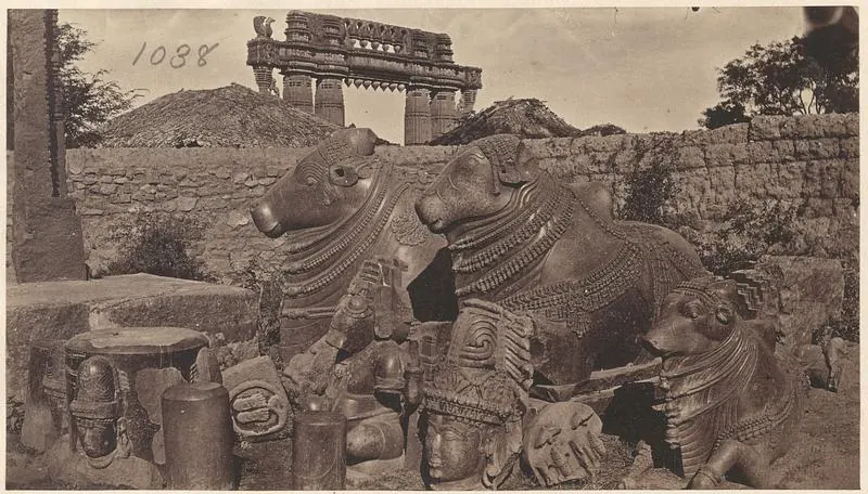 Two large carved stone bulls lying side by side among broken carved stones, a rough wall behind them and the top of a stone gateway rising above it.
