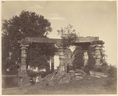 Four carved stone pillars holding up a broken beam among large trees, with fallen carved blocks on the ground and a man seated among them.