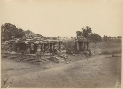 A low stone temple with a broken roof on a moulded platform in open ground, small trees growing on the roof and a small shrine to the right.
