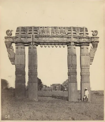 A tall stone gateway of four carved pillars and a carved crossbeam with stone swans at its ends, standing in open ground with a man beside the right pillar.