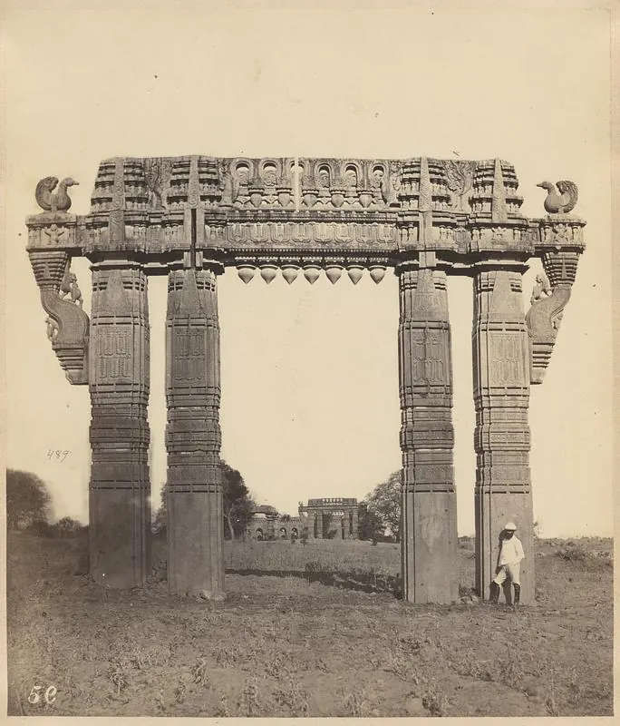 A tall stone gateway of four carved pillars and a carved crossbeam with stone swans at its ends, standing in open ground with a man beside the right pillar.