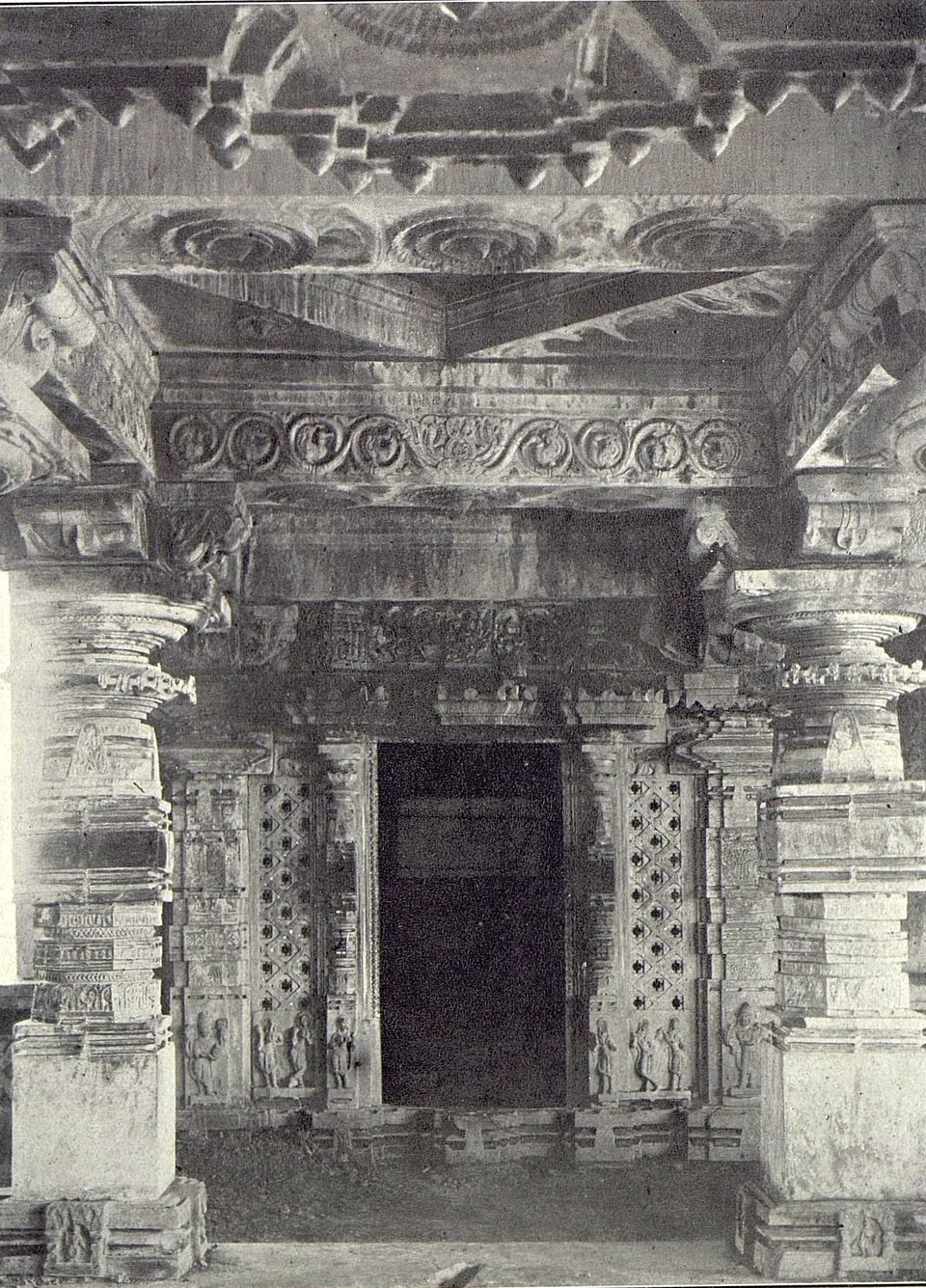Inside a temple hall: a dark doorway with pierced screens on both sides, under carved beams and a ceiling of projecting brackets, between carved pillars.