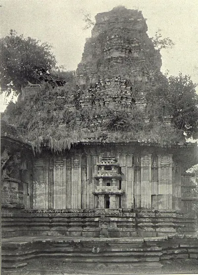 The tower over the shrine of the Ramappa temple, its upper tiers thick with grass and plants, above a plain wall with a small carved niche and a moulded base.