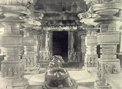 The interior of a small temple: a dark shrine doorway framed by carved jambs between heavy lathe-turned pillars, with a Nandi in the foreground.
