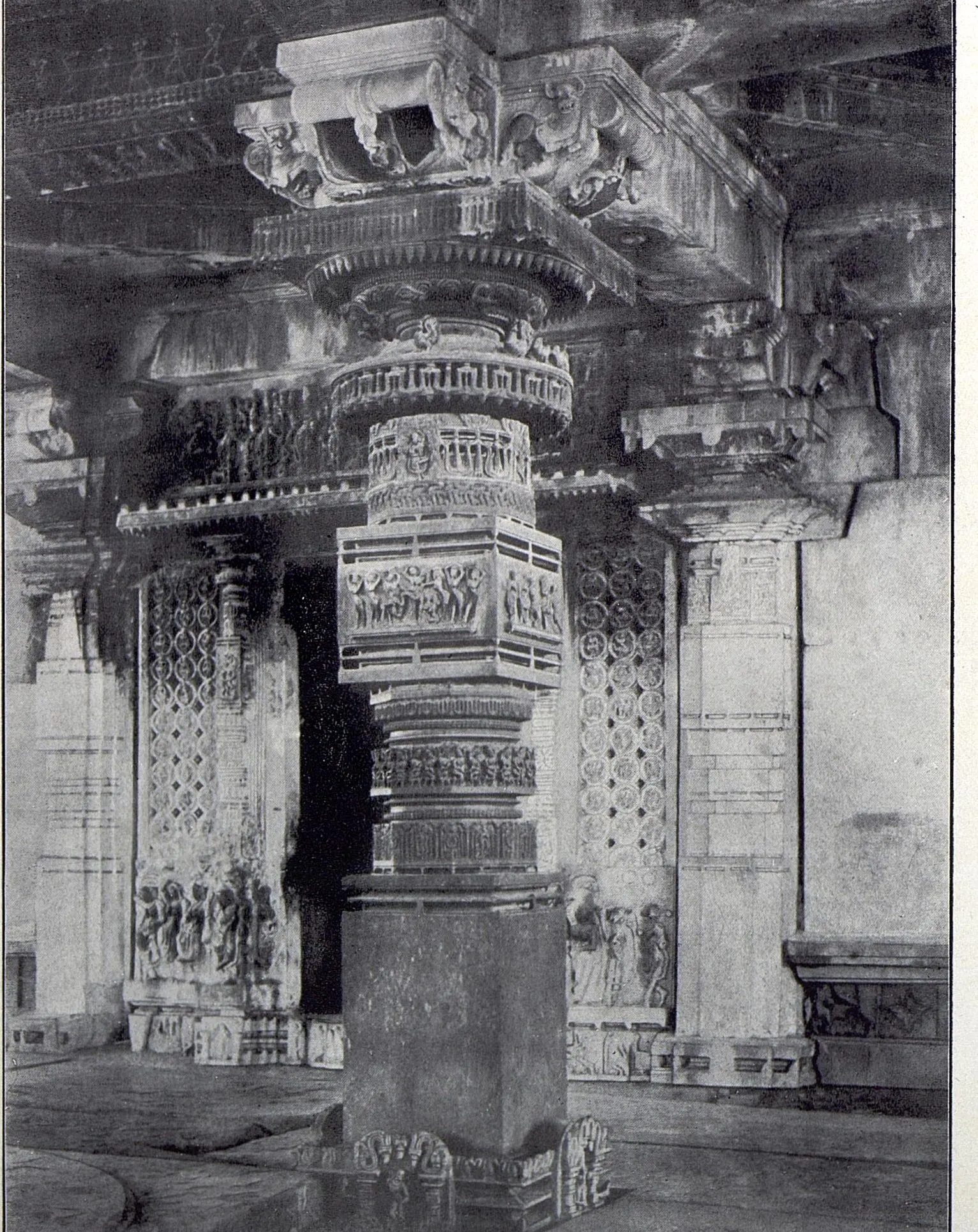 A polished stone pillar with a square base, a carved cubical block with figure panels and lathe-turned rings, under a bracket capital, with pierced screens and carved pilasters behind.