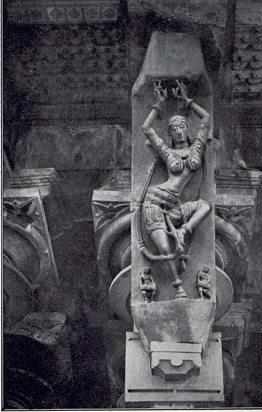 A slanting stone bracket carved with a dancer whose arms are raised above her head, with two small figures at her feet, above a pillar capital.