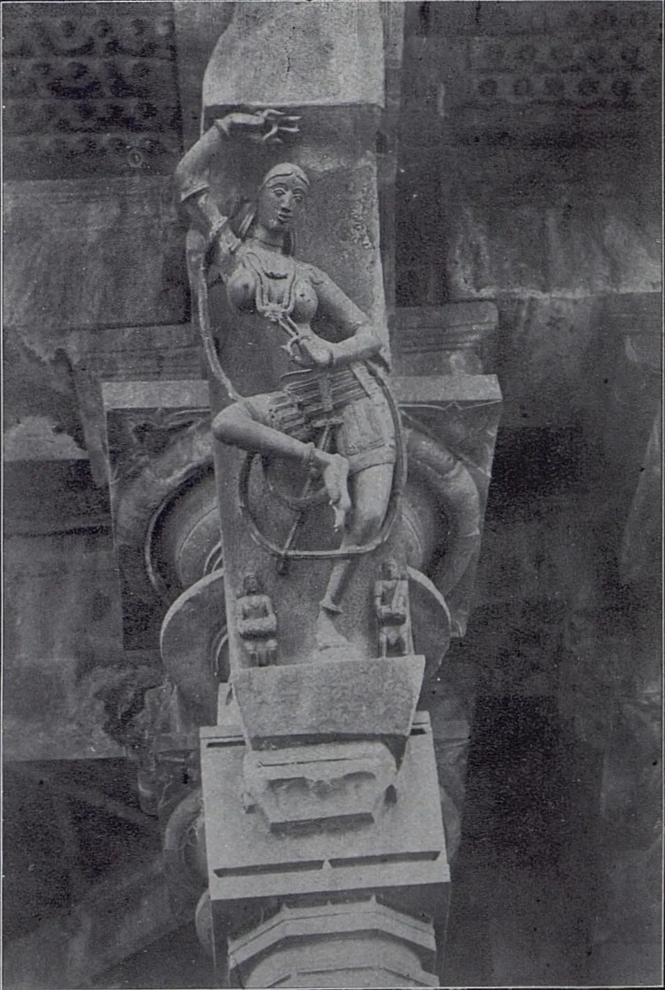 A slanting stone bracket carved with a dancer, one arm raised and a long looped cord or creeper around her body, with two small figures at her feet, above a pillar capital.