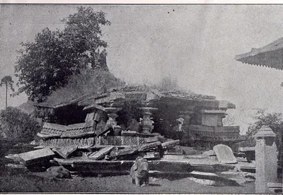 A collapsed stone temple on a stepped platform, its beams and carved blocks fallen, with a tree behind and the eave of another temple at the right edge.