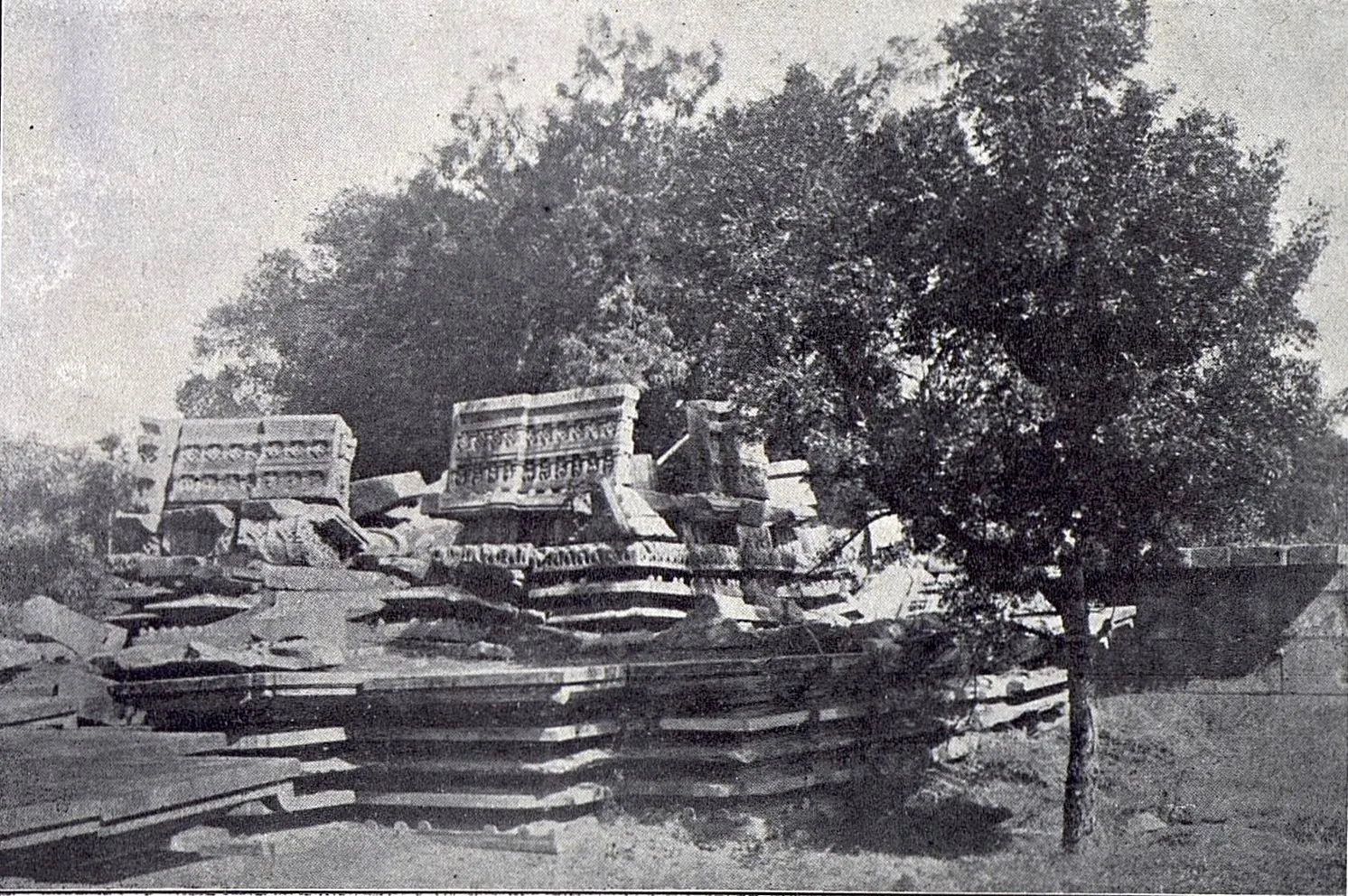 The fallen stones of a pavilion on a stepped platform, two carved roof blocks still standing on top, with a tree in front.