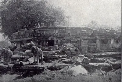 A ruined temple with low walls and a doorway behind heaps of fallen stone, two stone elephants in the foreground and a large tree behind.