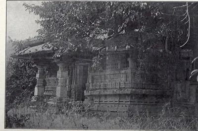 A small stone temple with a pillared porch and carved walls, half hidden by trees and tall grass.
