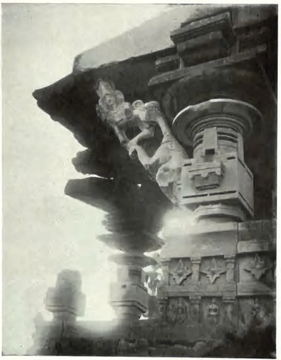 A dark stone bracket figure of a woman standing against a slanting bracket under the eave of the main Ghanpur temple, above a carved pillar capital and a moulded wall, lit from the side.