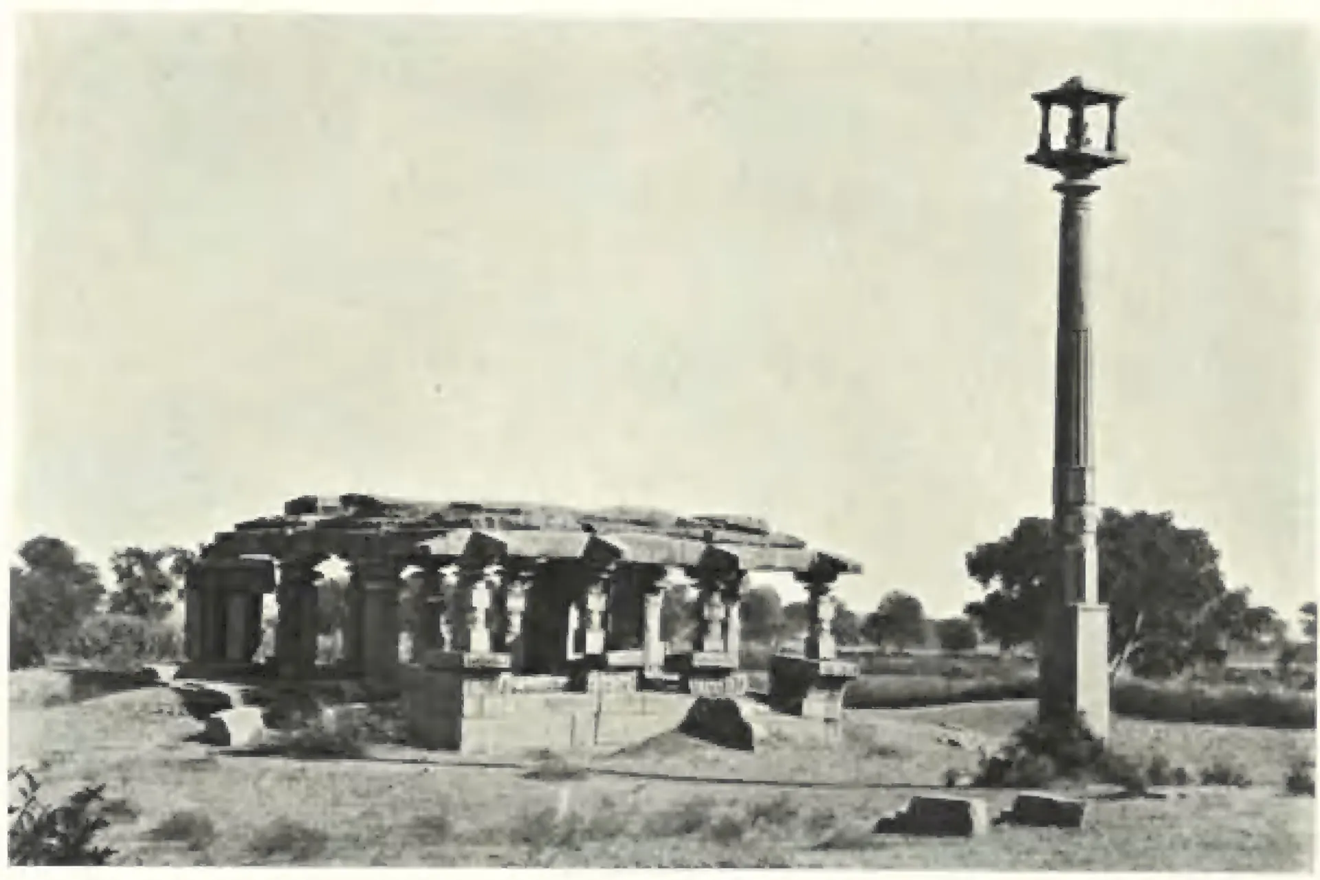 A ruined pillared temple with a flat roof in open ground, beside a tall monolithic pillar crowned by a small pavilion that once held a Garuda figure.