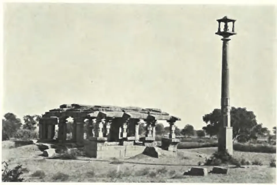 A ruined pillared temple with a flat roof in open ground, beside a tall monolithic pillar crowned by a small pavilion that once held a Garuda figure.