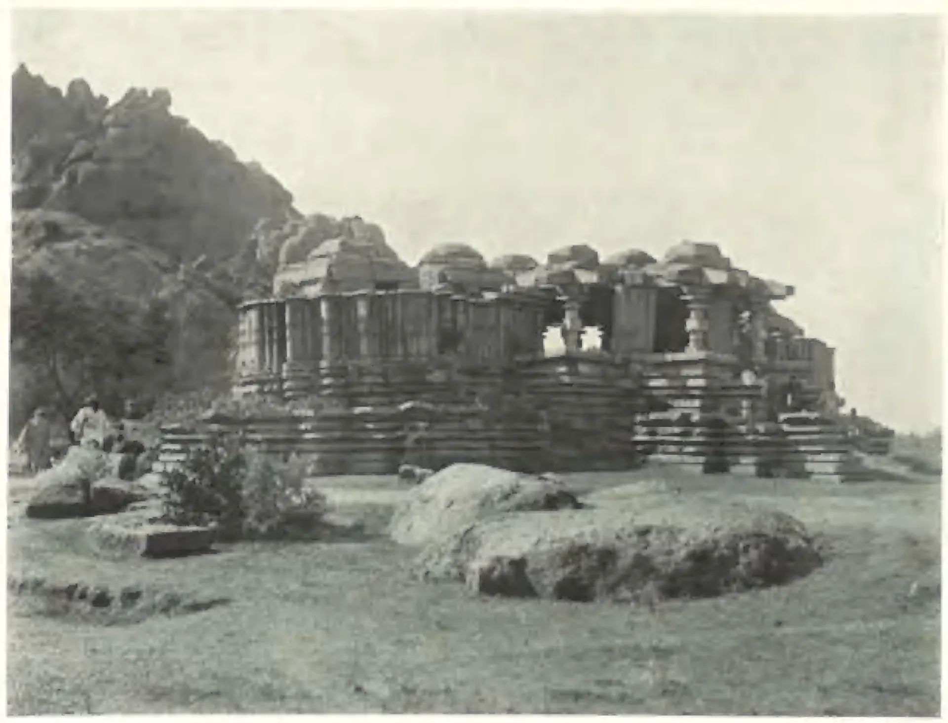 A large ruined triple-shrined stone temple on a moulded platform below a rocky hill, its shrines without towers, with boulders and two men in the foreground.
