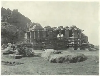 A large ruined triple-shrined stone temple on a moulded platform below a rocky hill, its shrines without towers, with boulders and two men in the foreground.