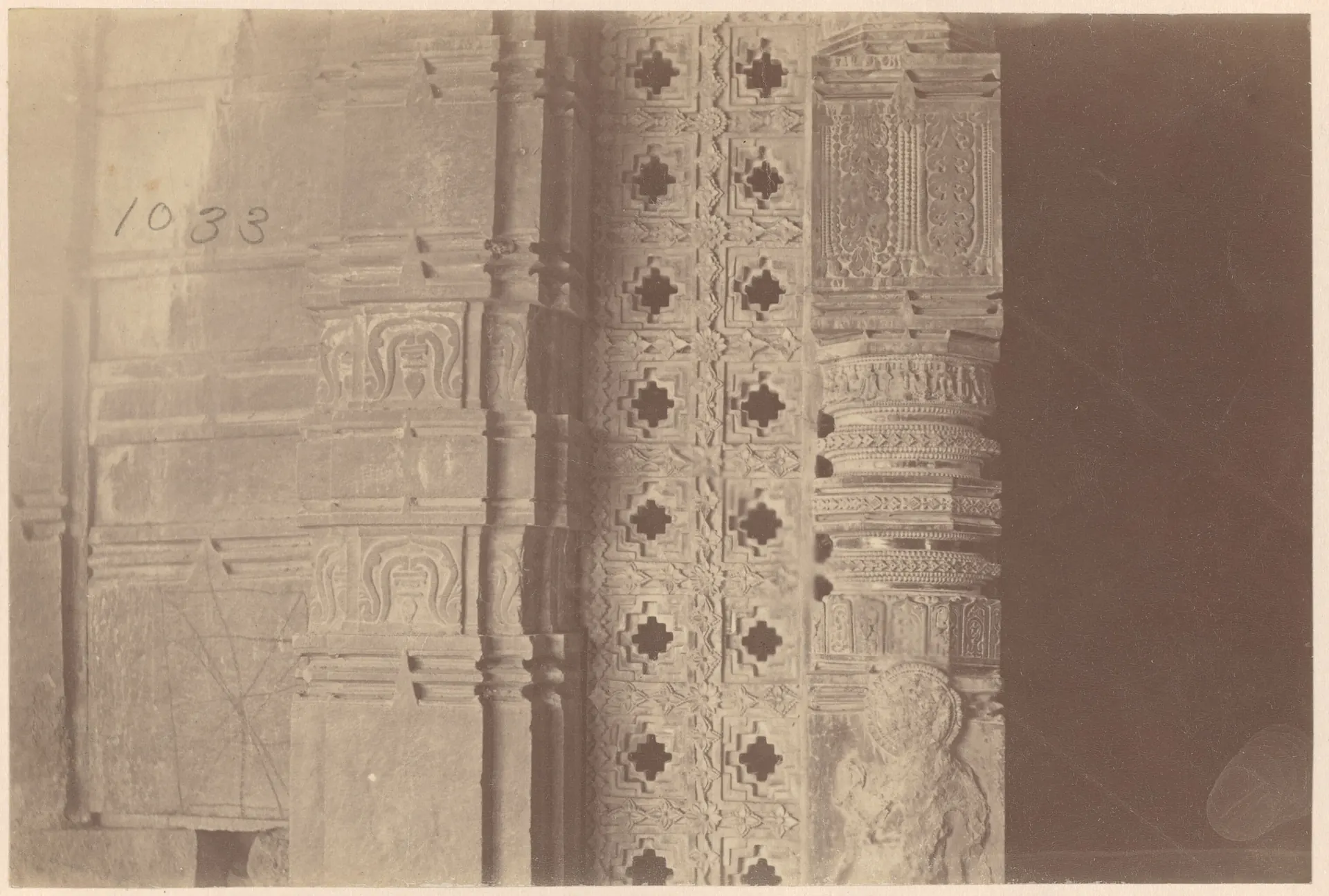 Close view of a temple doorway: a carved wall pilaster at the left, a stone screen pierced with stepped cross-shaped openings in the middle, and a turned, banded pillar and dark doorway at the right.