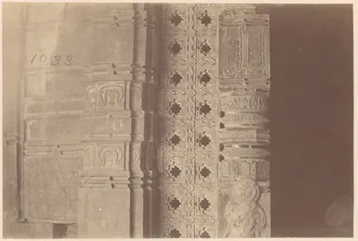Close view of a temple doorway: a carved wall pilaster at the left, a stone screen pierced with stepped cross-shaped openings in the middle, and a turned, banded pillar and dark doorway at the right.