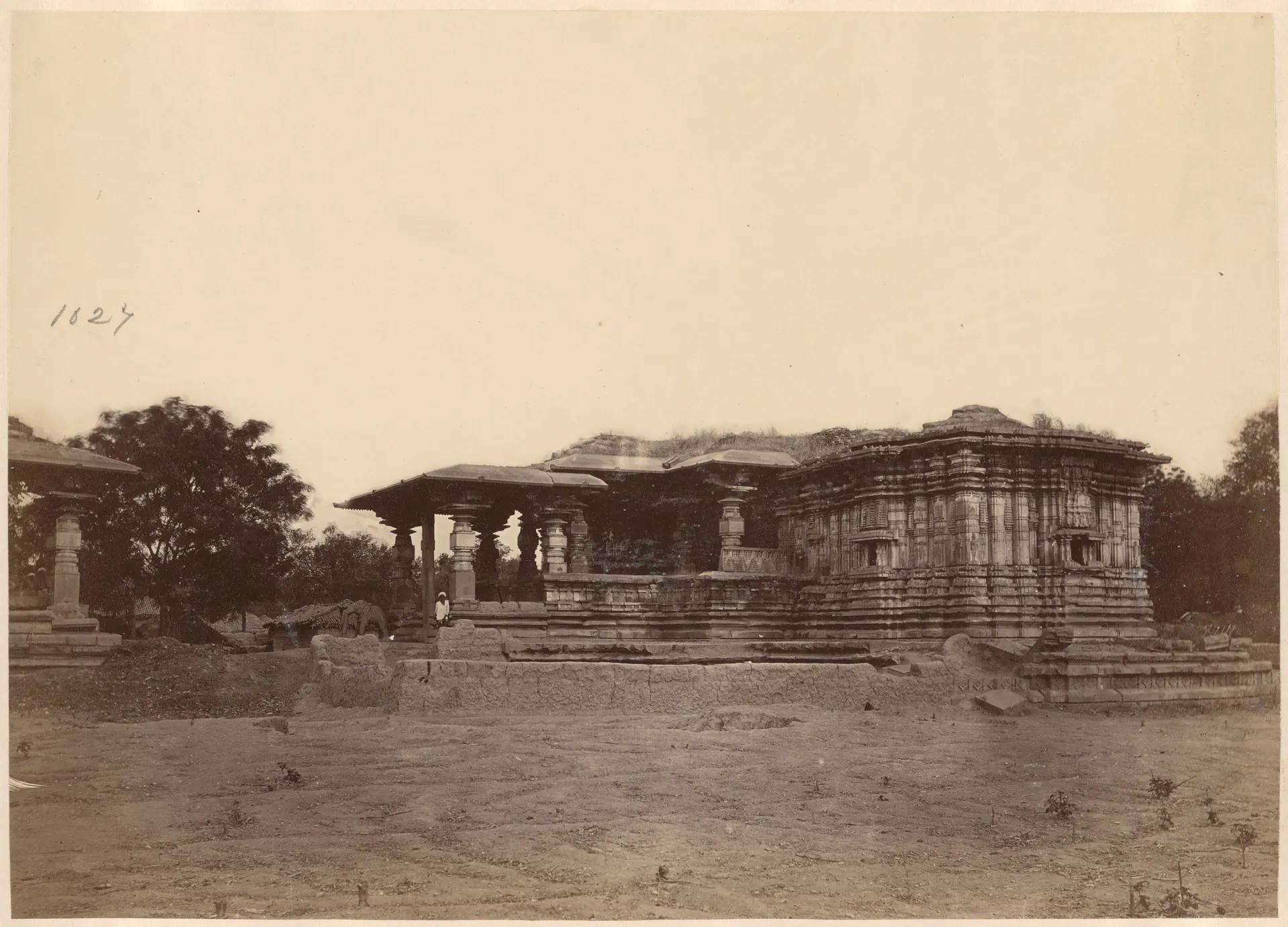 The Thousand Pillar Temple seen side-on across bare ground: a low pillared porch at the left of the carved, roofless main block on its stepped platform, a detached pavilion at the far left and trees behind.
