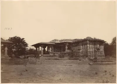 The Thousand Pillar Temple seen side-on across bare ground: a low pillared porch at the left of the carved, roofless main block on its stepped platform, a detached pavilion at the far left and trees behind.